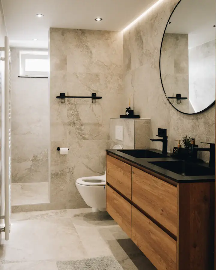 Salle d’eau rénovée : meuble vasque en bois, miroir rond et carrelage effet pierre